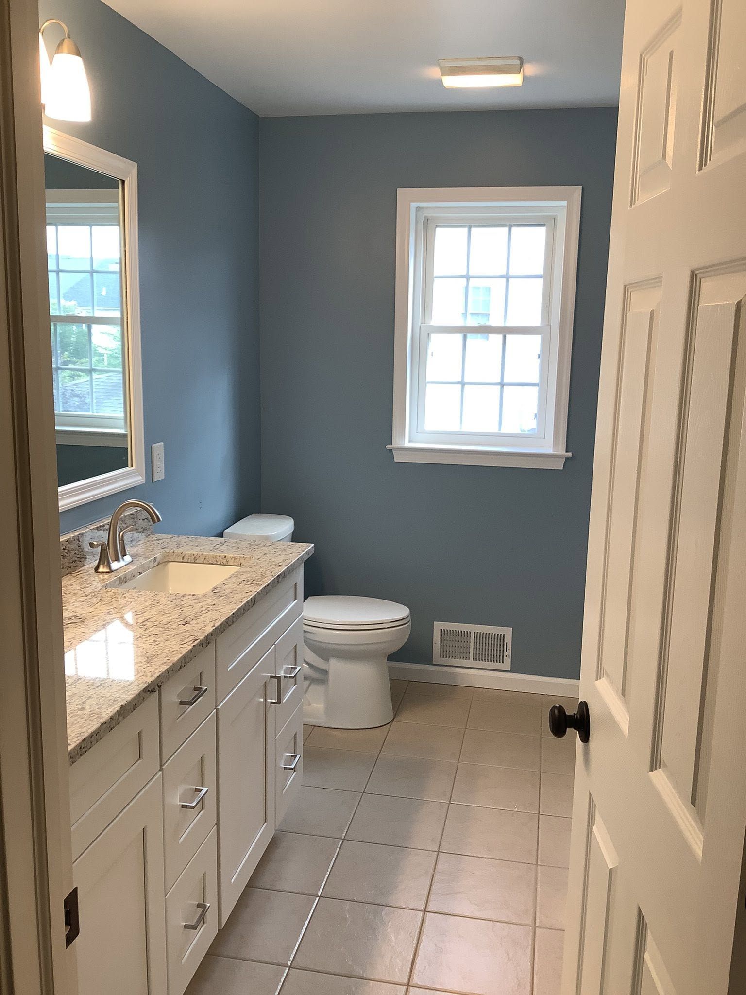 Narrow bathroom with blue walls, white vanity, toilet, window, and tiled floor, seen from the doorway