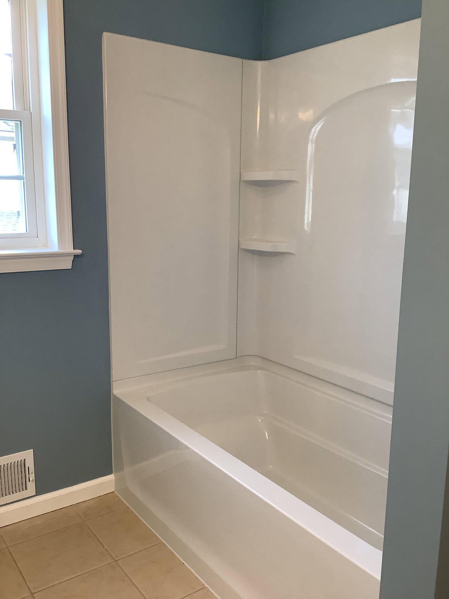 White bathtub and shower surround in a blue bathroom with a window and tiled floor