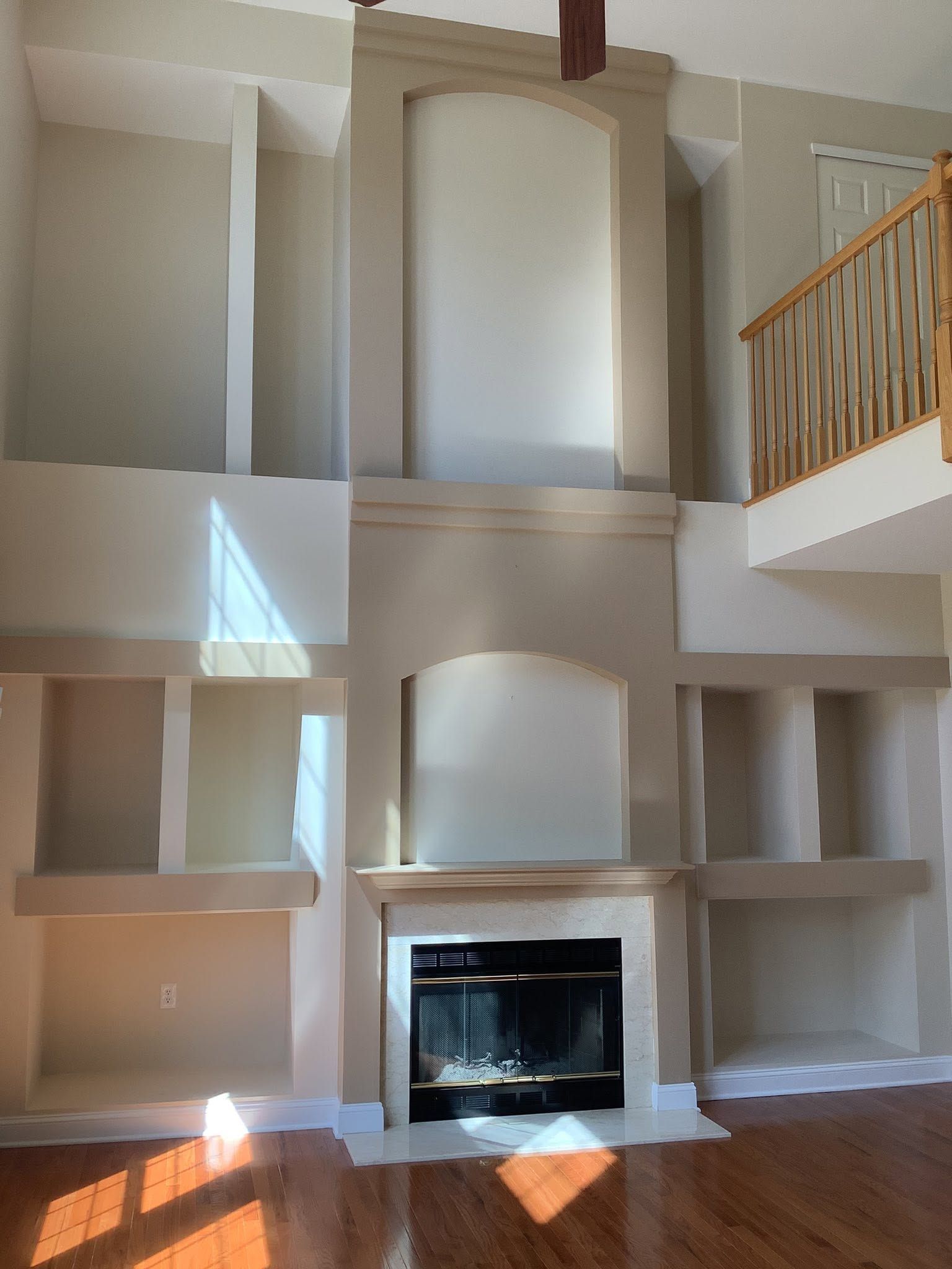 Living room wall with built-in shelves, a fireplace, and a staircase railing above; sunlight falls across the wall.