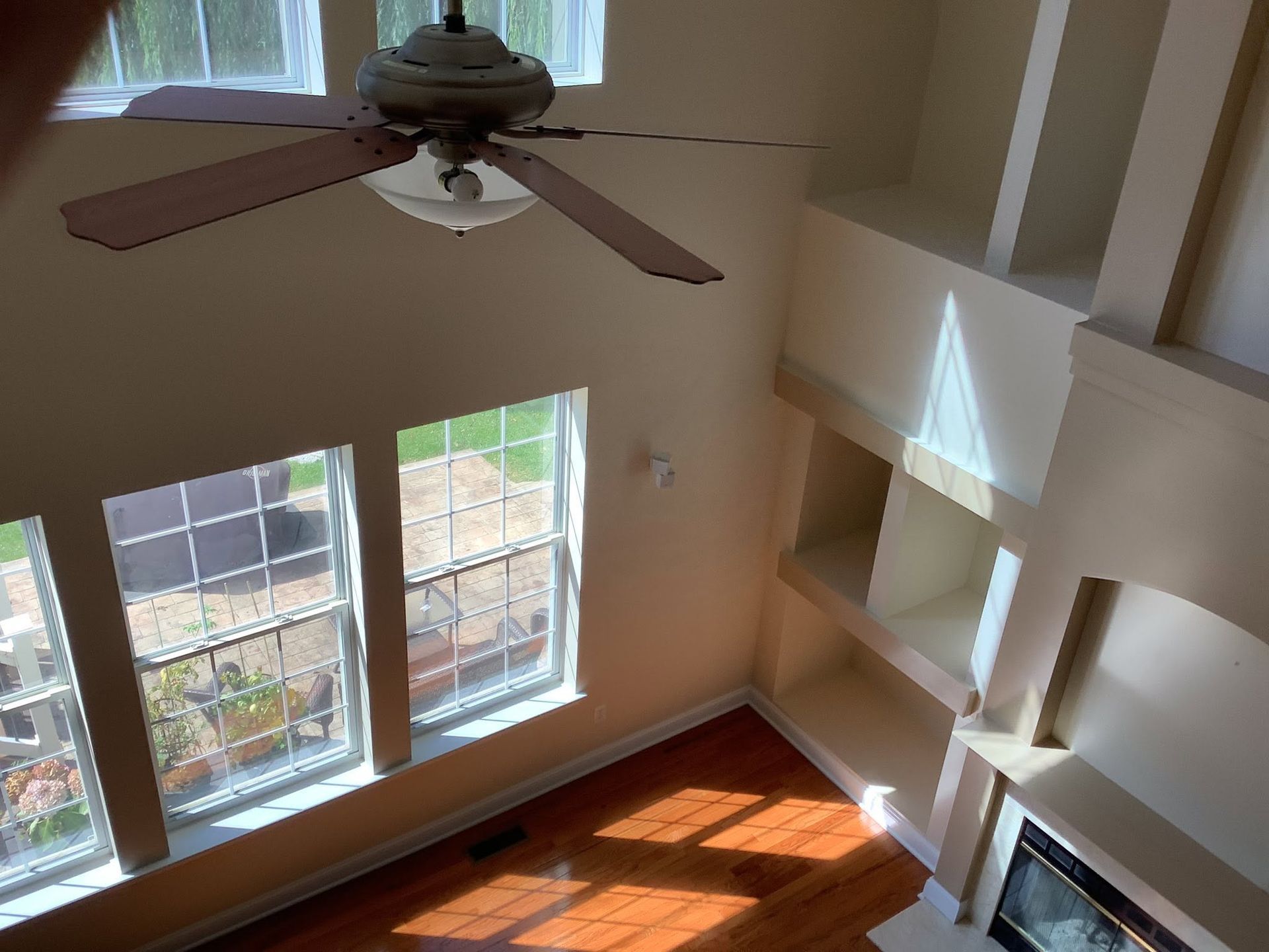 Empty living room with tall windows, ceiling fan, and sunlight streaming onto wood floors
