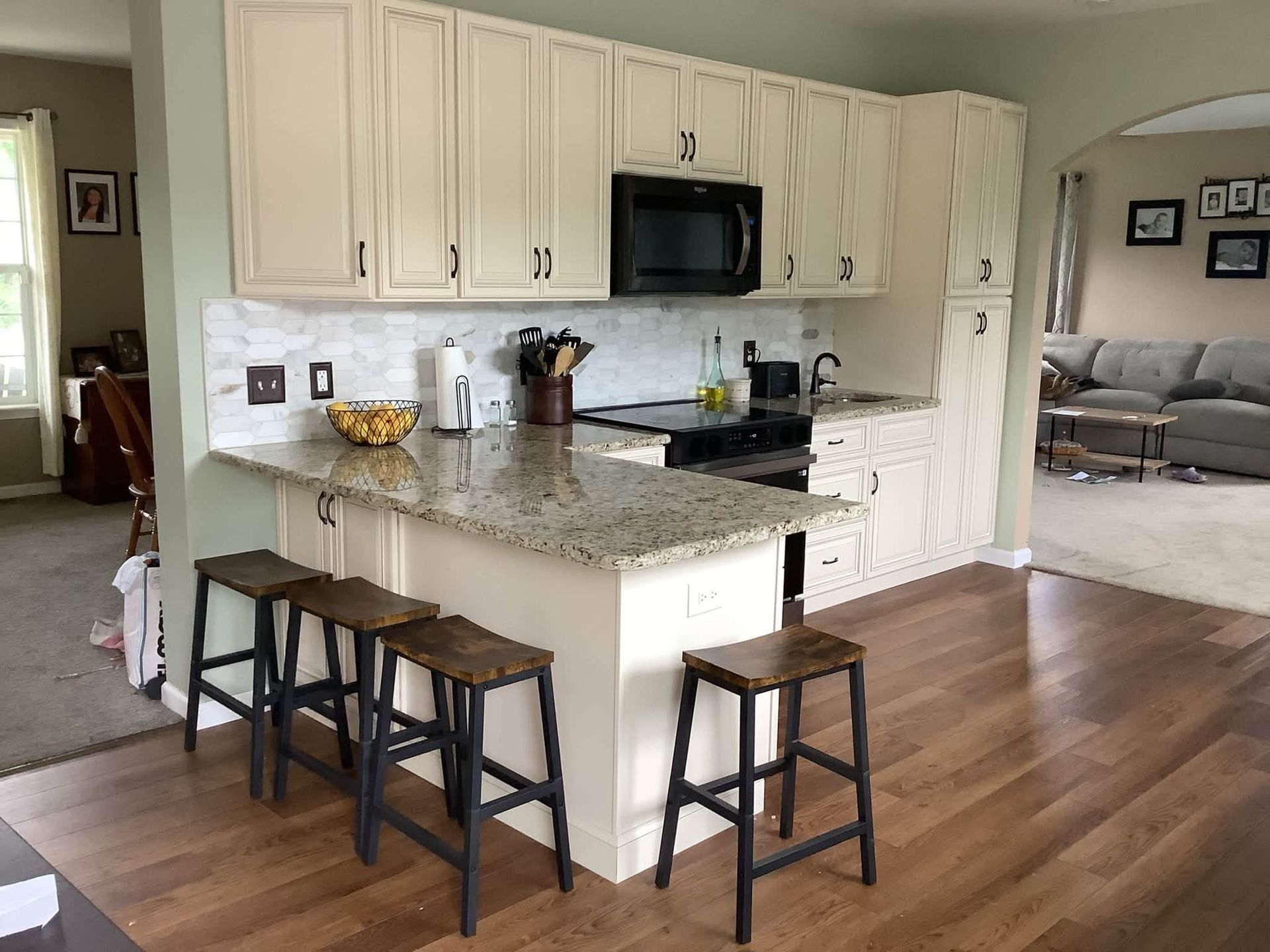 Kitchen with white cabinets, granite island, stools, and hardwood floors leading to a living room.