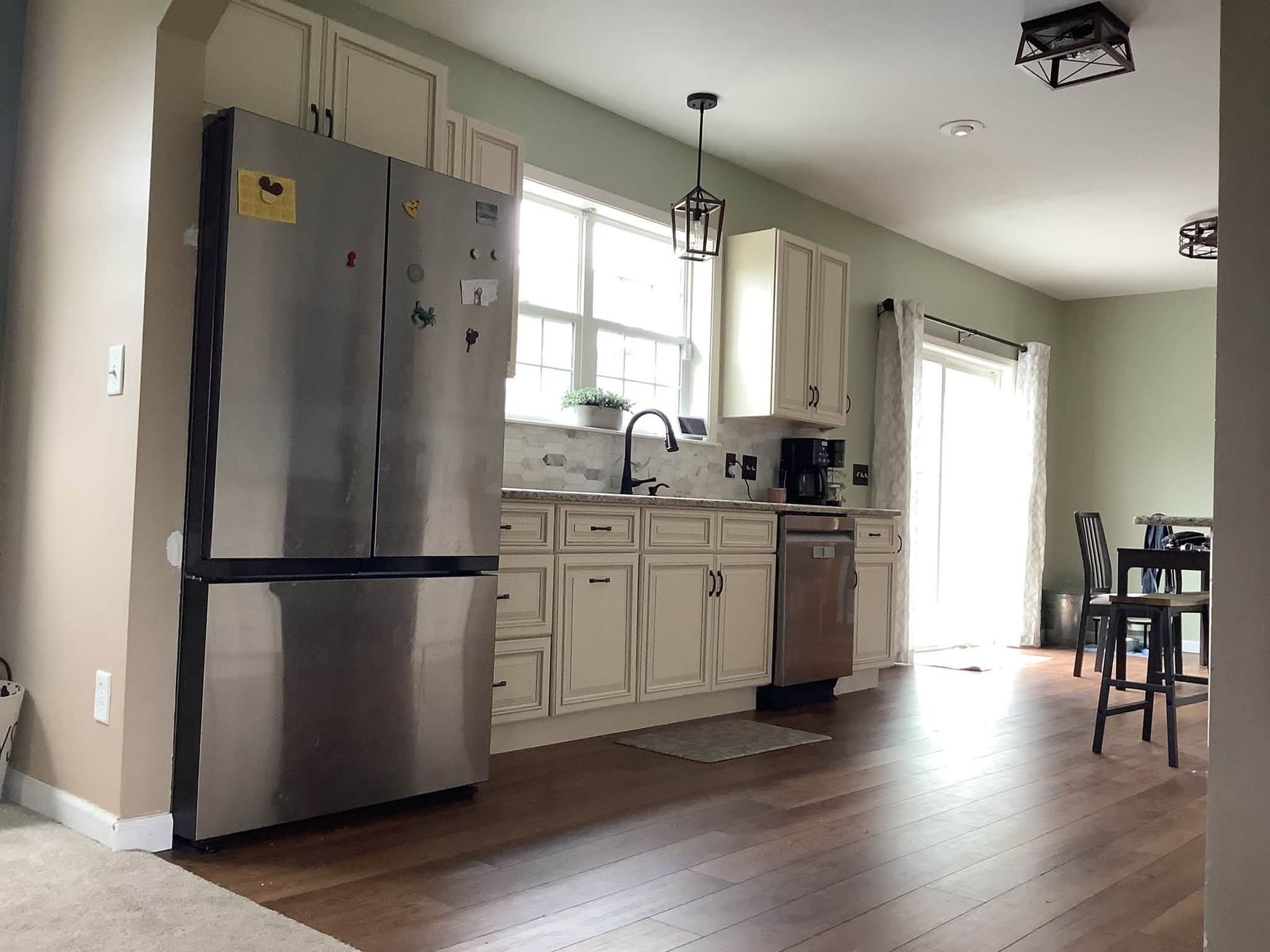 Bright kitchen with stainless steel fridge, white cabinets, window, and wood floor, opening to a dining area