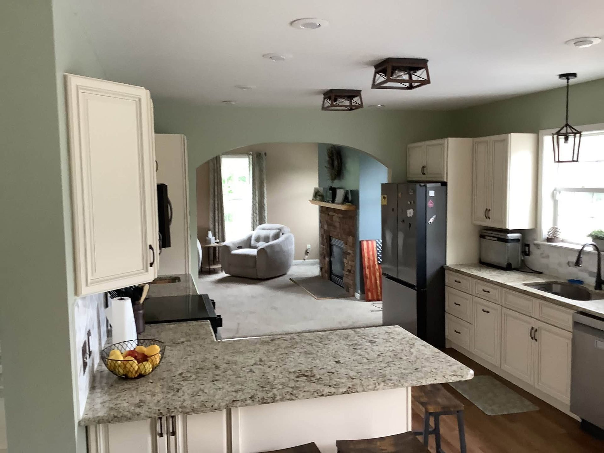 Bright kitchen with granite island, white cabinets, stainless fridge, and a view into a living room.
