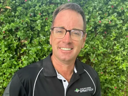Jeremy Jones wearing glasses and a black shirt is smiling in front of a hedge.