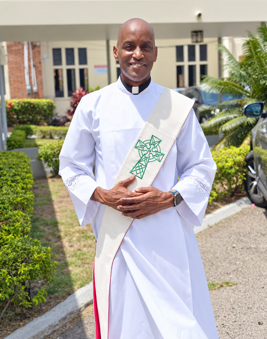 Clergyman with dark skin wearing white robe and green sash with cross necklace, standing indoors.