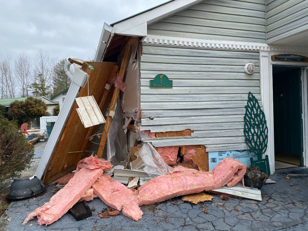 Damaged house exterior. Wall collapsed, insulation visible. Gray siding, green door, overcast sky.