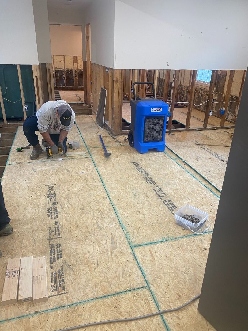 Man cuts wood flooring in a room under construction, with a blue dehumidifier and exposed wall studs visible.