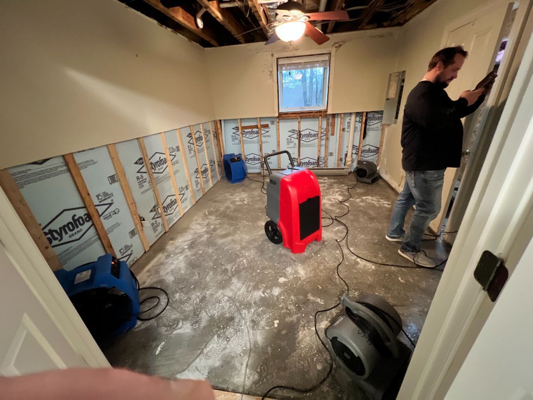 Man working on a room with moisture damage, removing drywall. Dehumidifiers and fans running, insulation visible.