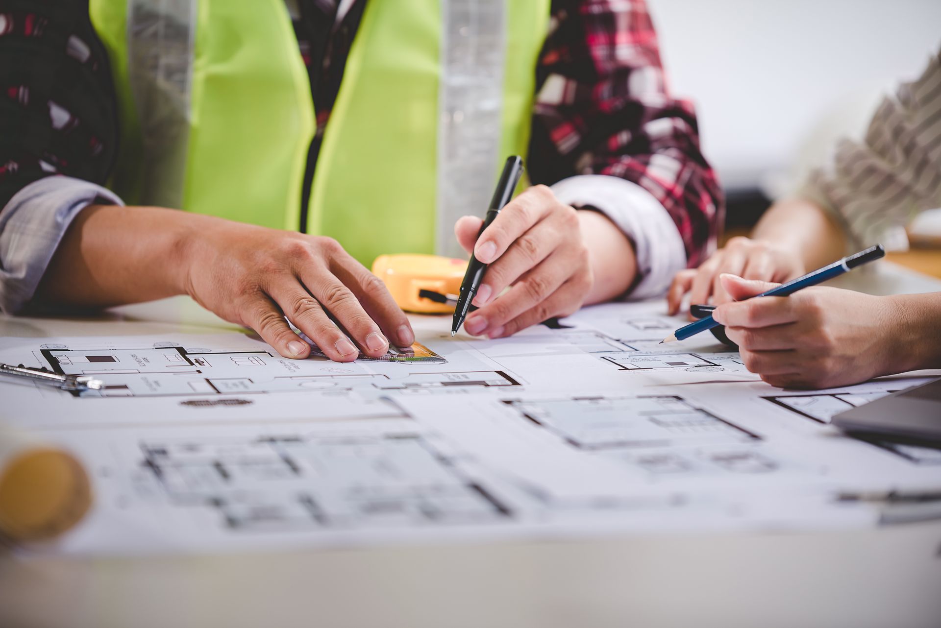 Hands using pens and rulers on architectural plans. A person in a safety vest.