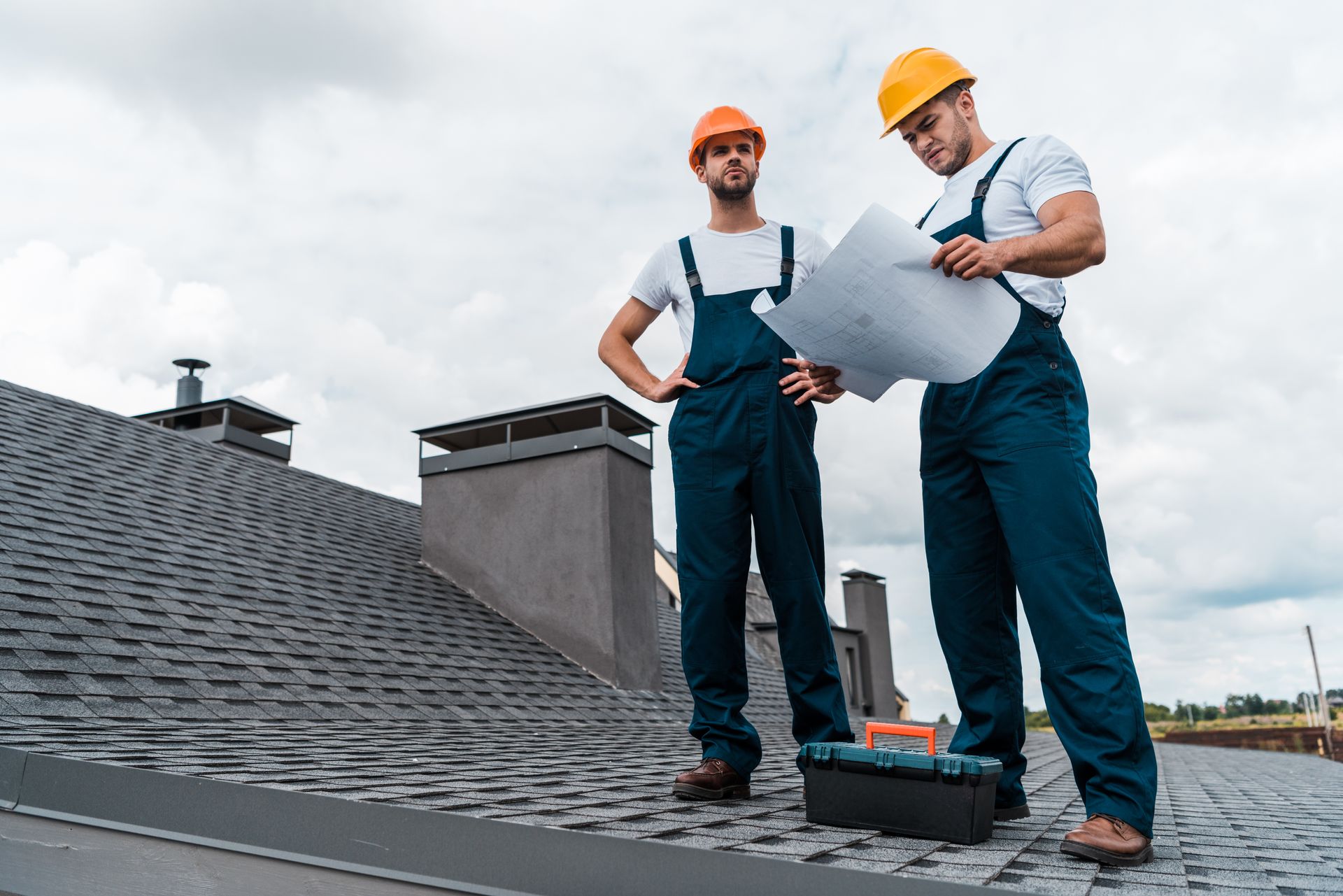 Two roofers in orange and yellow hard hats reviewing blueprints on a shingled roof.