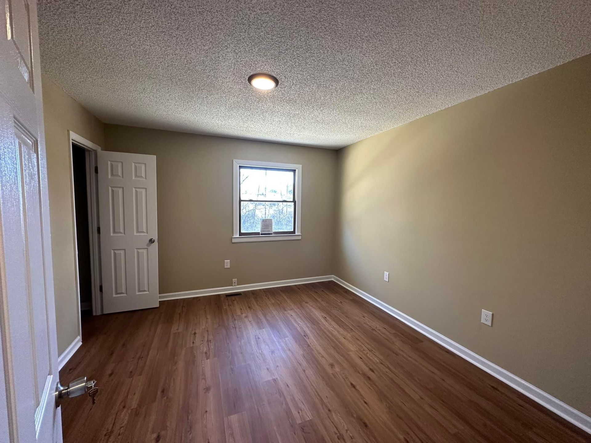Empty room with tan walls, brown wood-look floor, window, and white door.