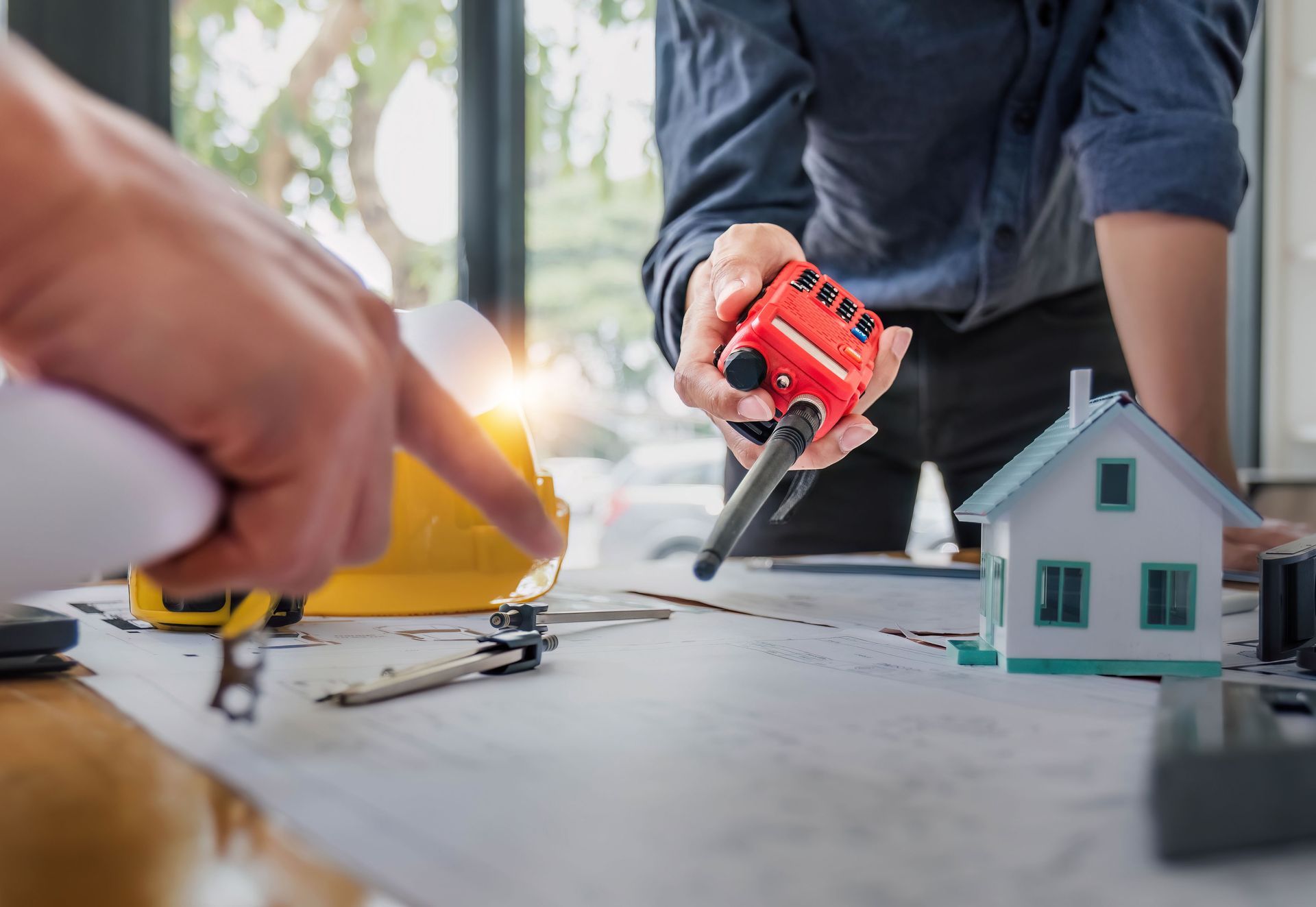 Hands and tools building a miniature house model on a table with a red toy vehicle nearby