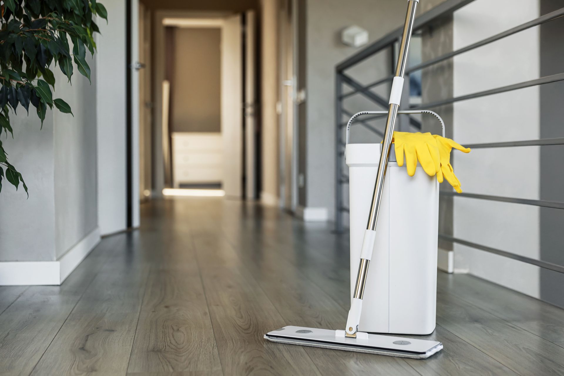 Mop and bucket with yellow gloves rest on a wooden floor in a hallway near a railing. Mop and bucket with yellow gloves rest on a wooden floor in a hallway near a railing.
