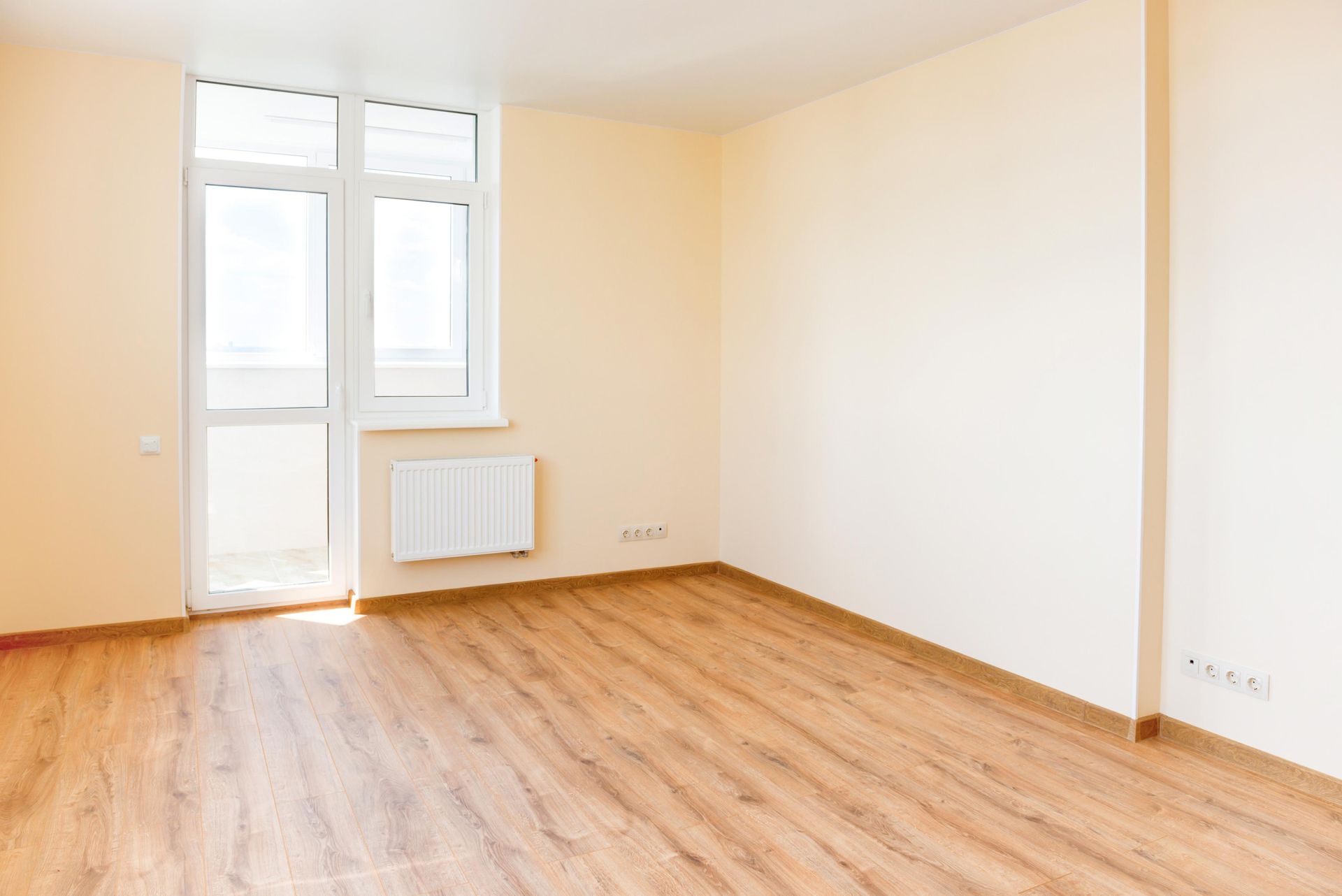 Empty room with wood-look floor, window/door to balcony, beige walls, and white radiator.