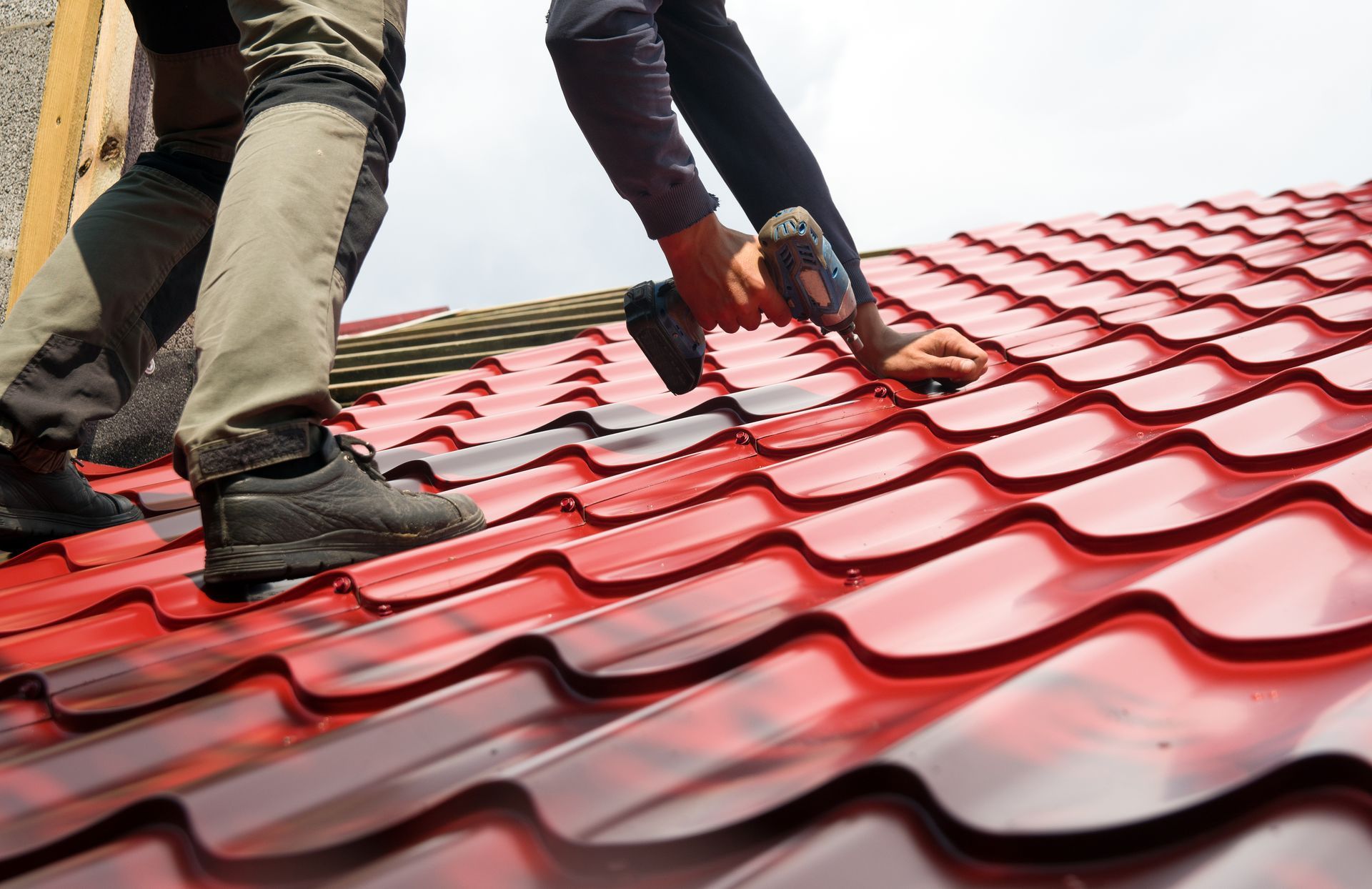 Two workers installing red metal roofing with a drill.