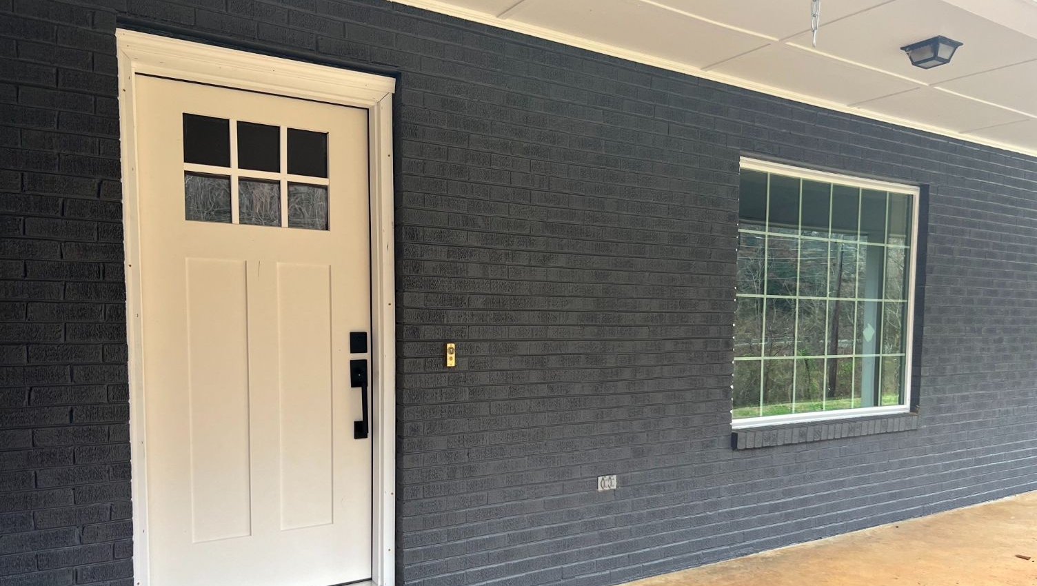 A white, six-paneled front door next to a window on a dark, textured brick wall under a covered porch.