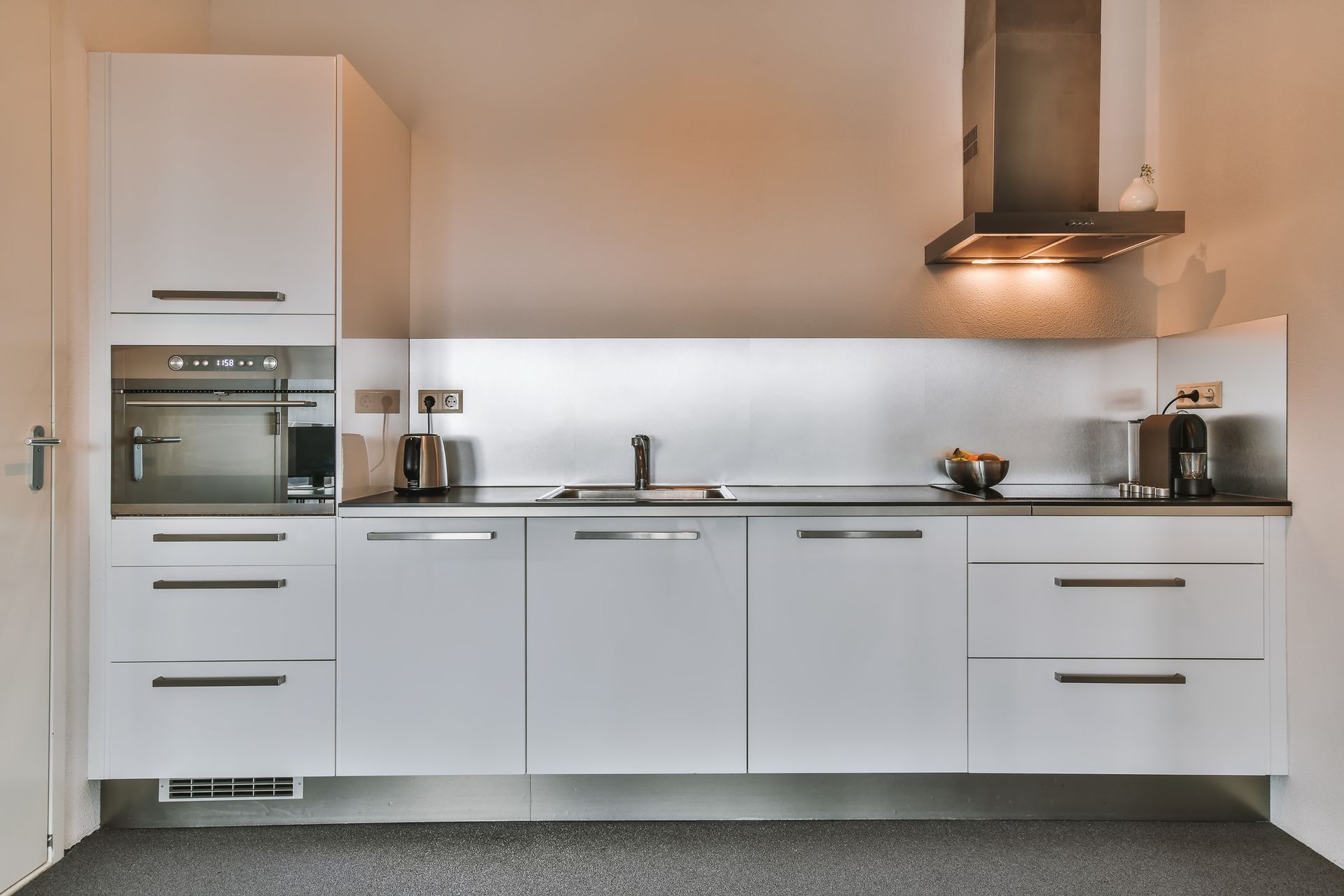 White modern kitchen with cabinets, oven, and sink against a neutral wall; a range hood above the cooktop.