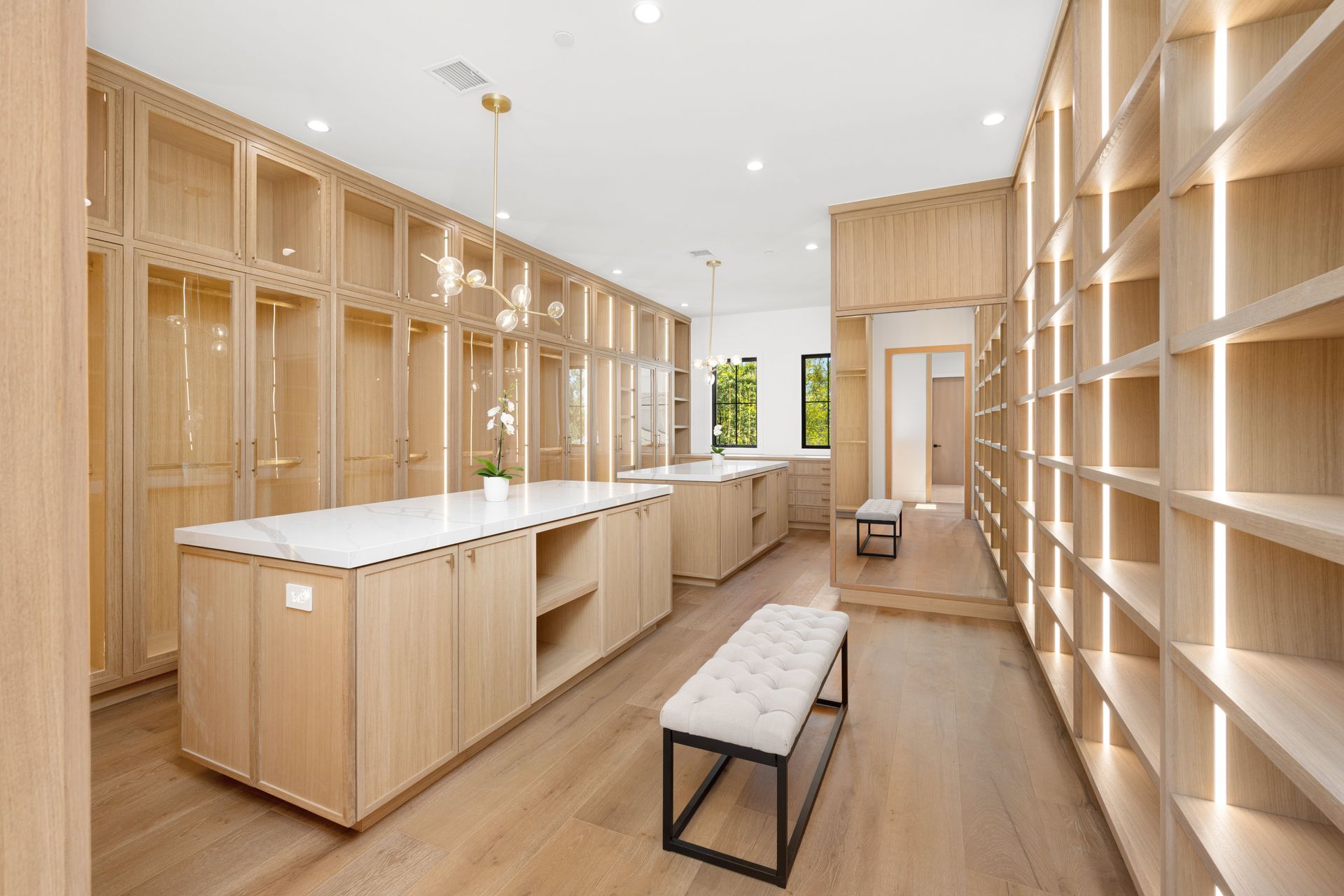 Spacious, light wood walk-in closet with shelves, cabinets, and islands. Bench and chandelier add elegance.