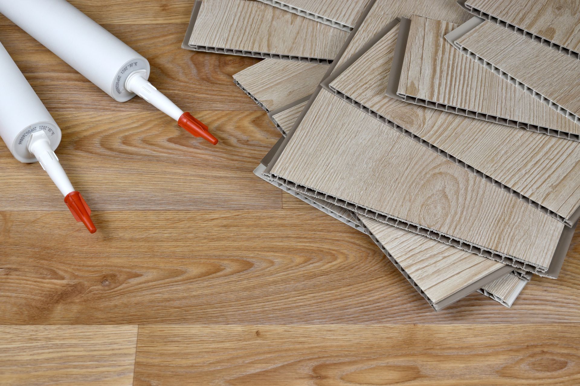 Two caulk tubes and wood-look panels on a hardwood floor.