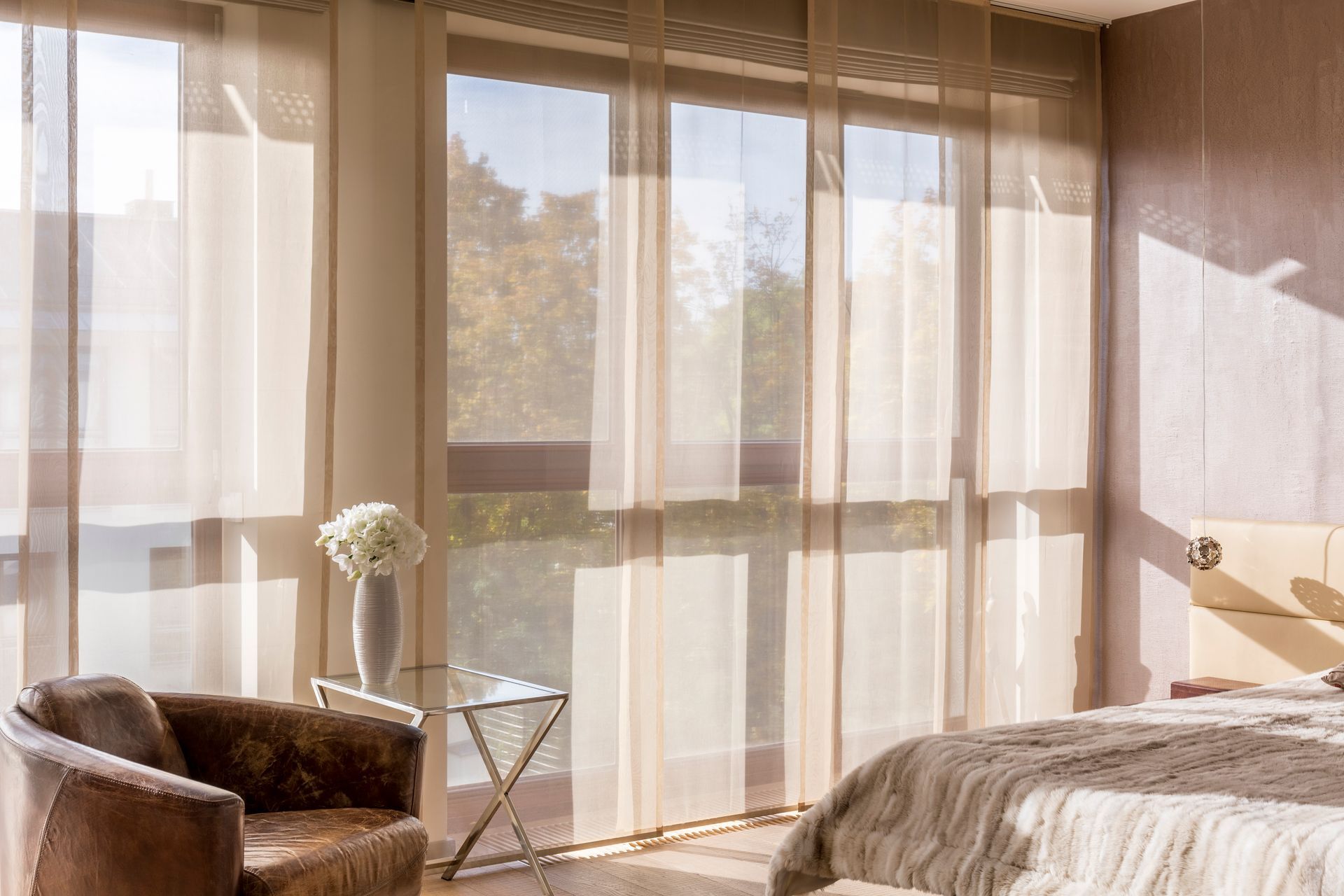 Room with sheer curtains, a chair, side table, and bed. Sunlight streams through the windows.