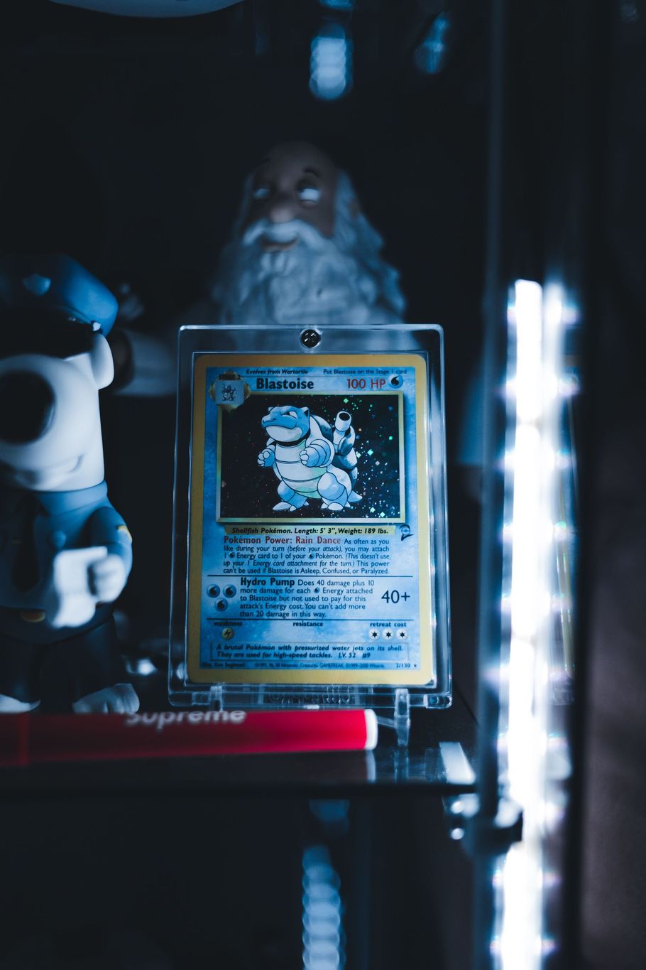 Holographic trading card in a display case, lit blue, with a blurred figurine and figure in the background