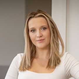 A woman with long blonde hair is wearing a white shirt and looking at the camera.