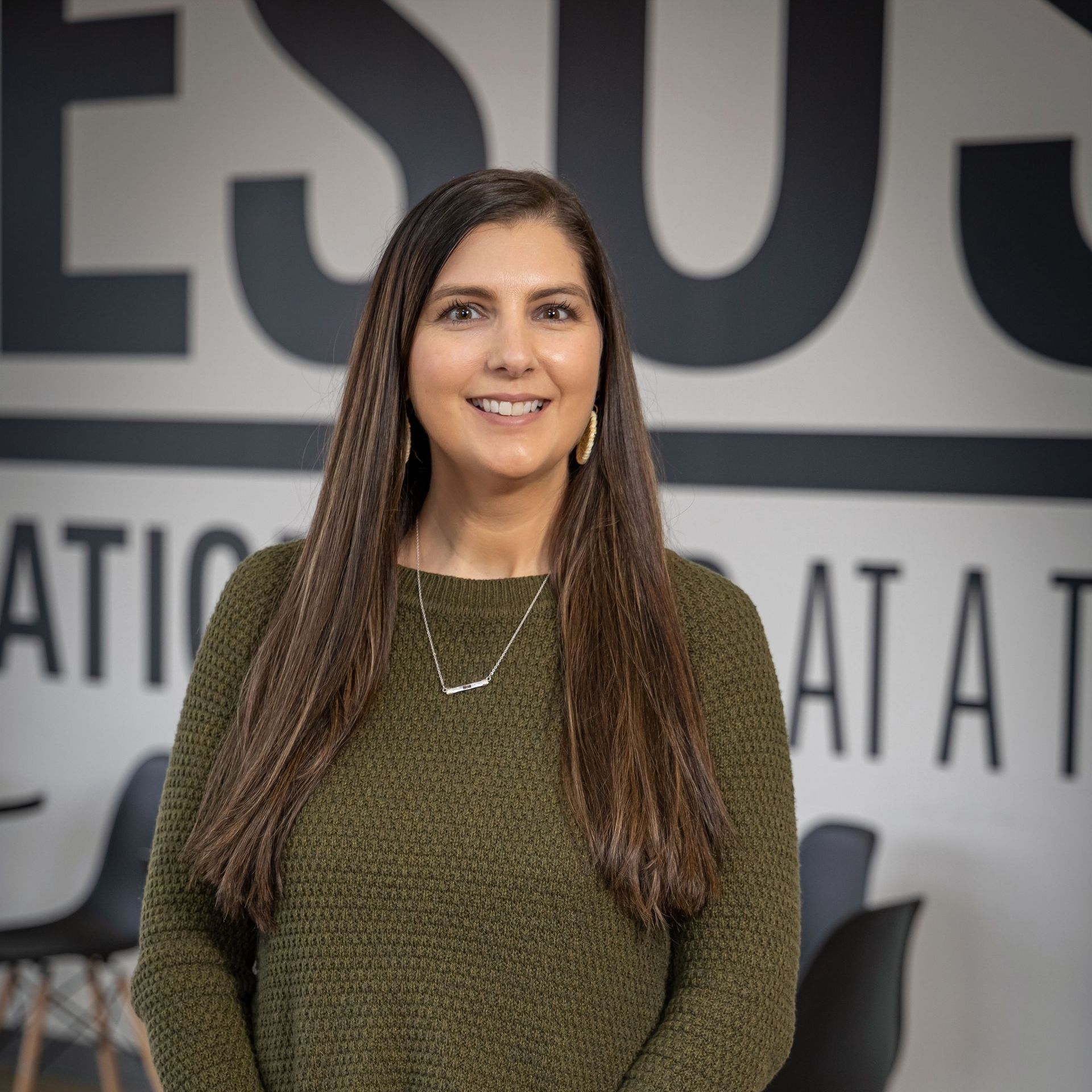 A woman in a green sweater is smiling in front of a sign that says jesus