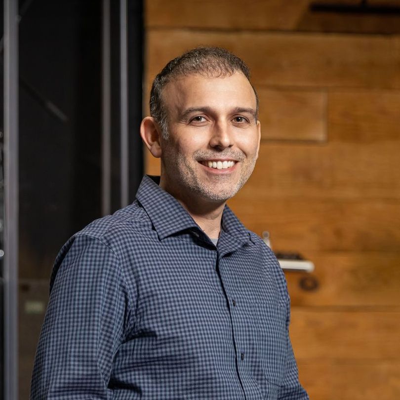A man in a blue shirt is smiling for the camera while standing in front of a wooden wall.