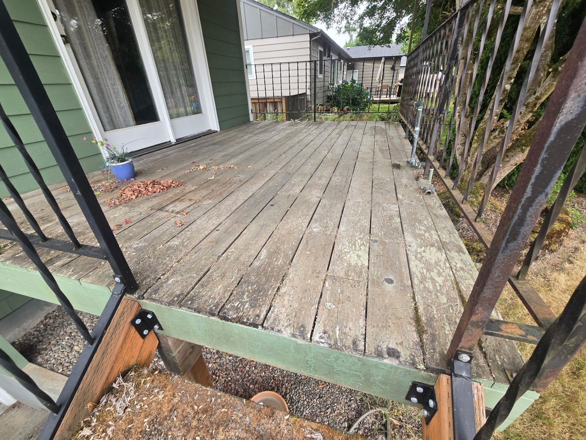 Weathered wooden backyard deck beside a green house, with black railings and trees in the background.