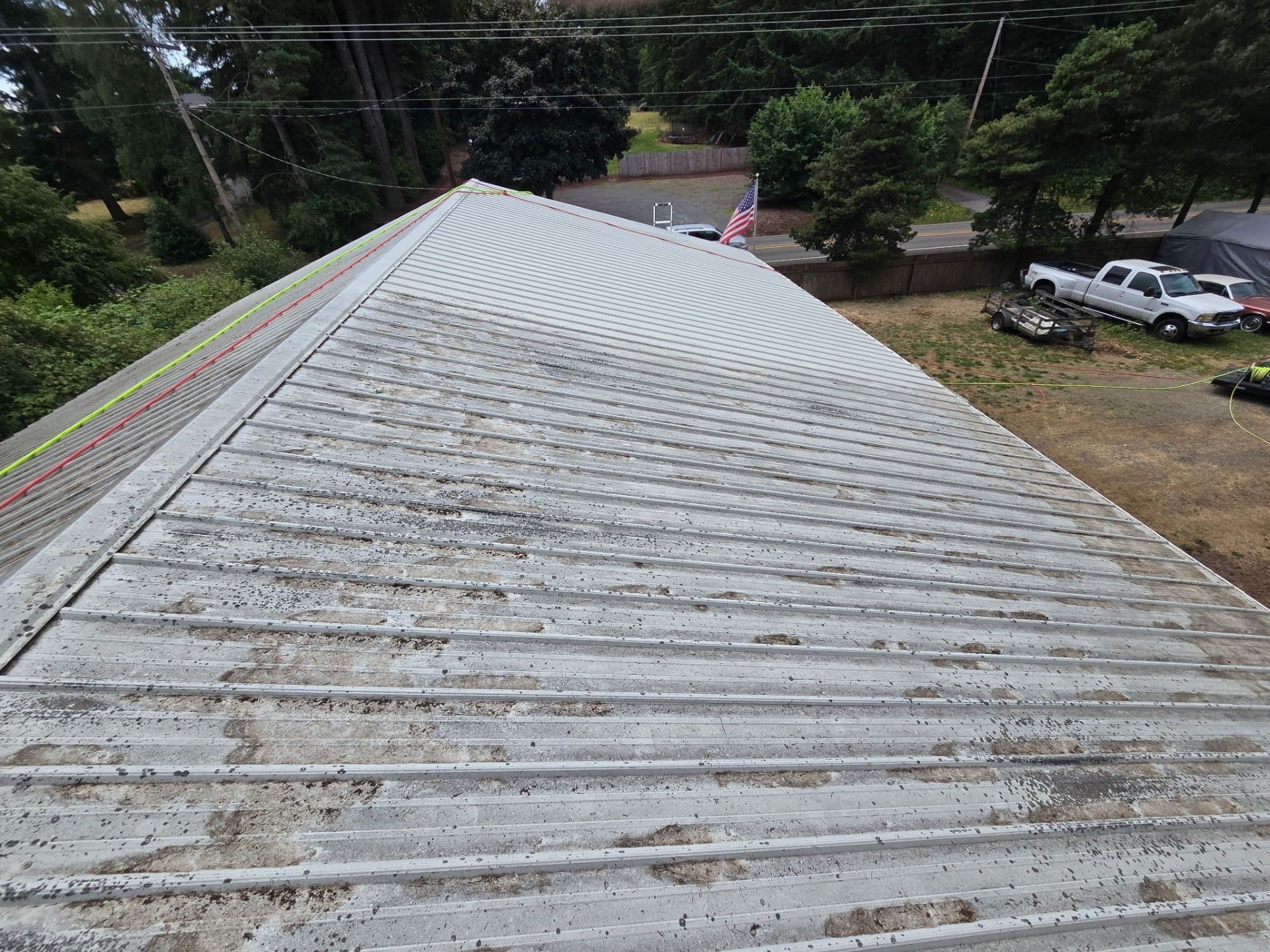 Roof viewed from above with weathered gray metal panels and trees, cars, and yard below
