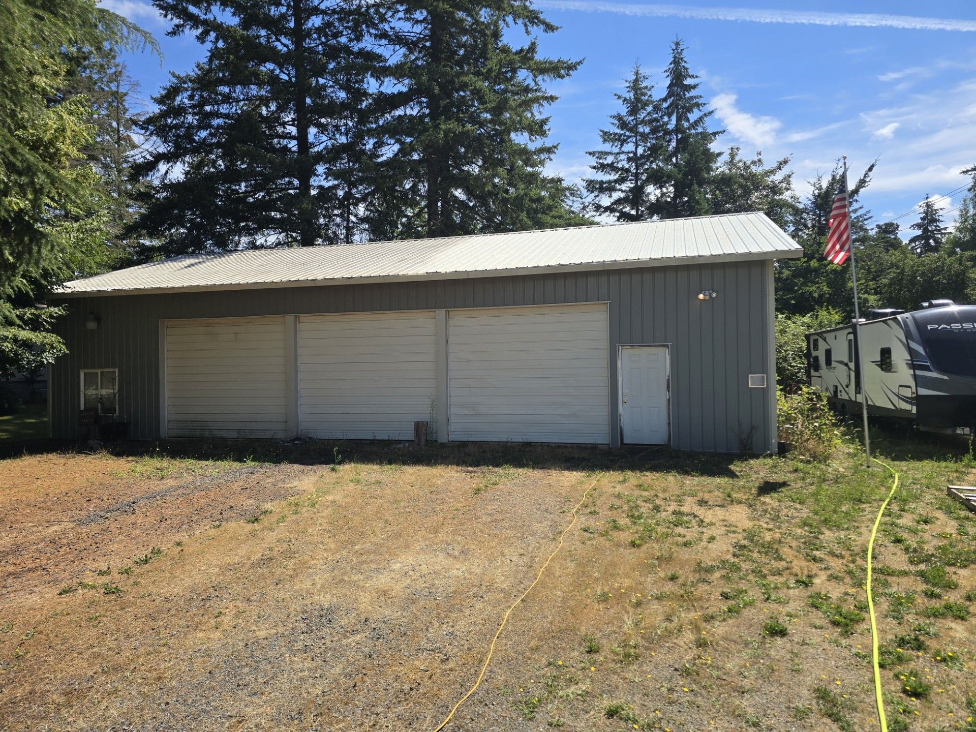 Gray metal garage with three white doors, a side door, and an American flag on a sunny rural lot.