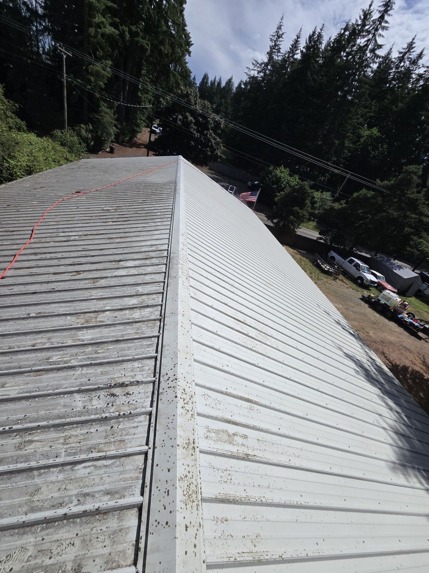Corrugated metal roof with white and weathered gray panels, viewed from the ridge beside trees and parked cars