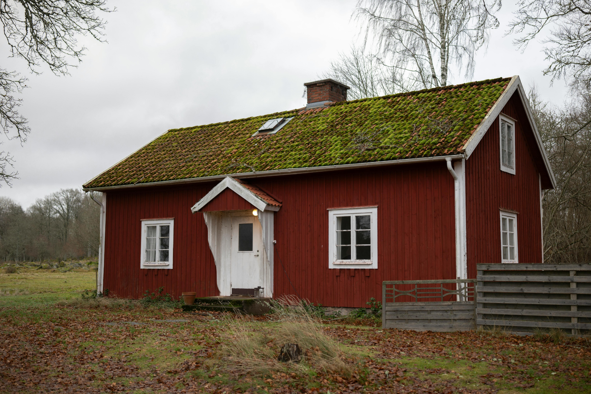 Red wooden cottage with a mossy roof in a rural field under gray skies