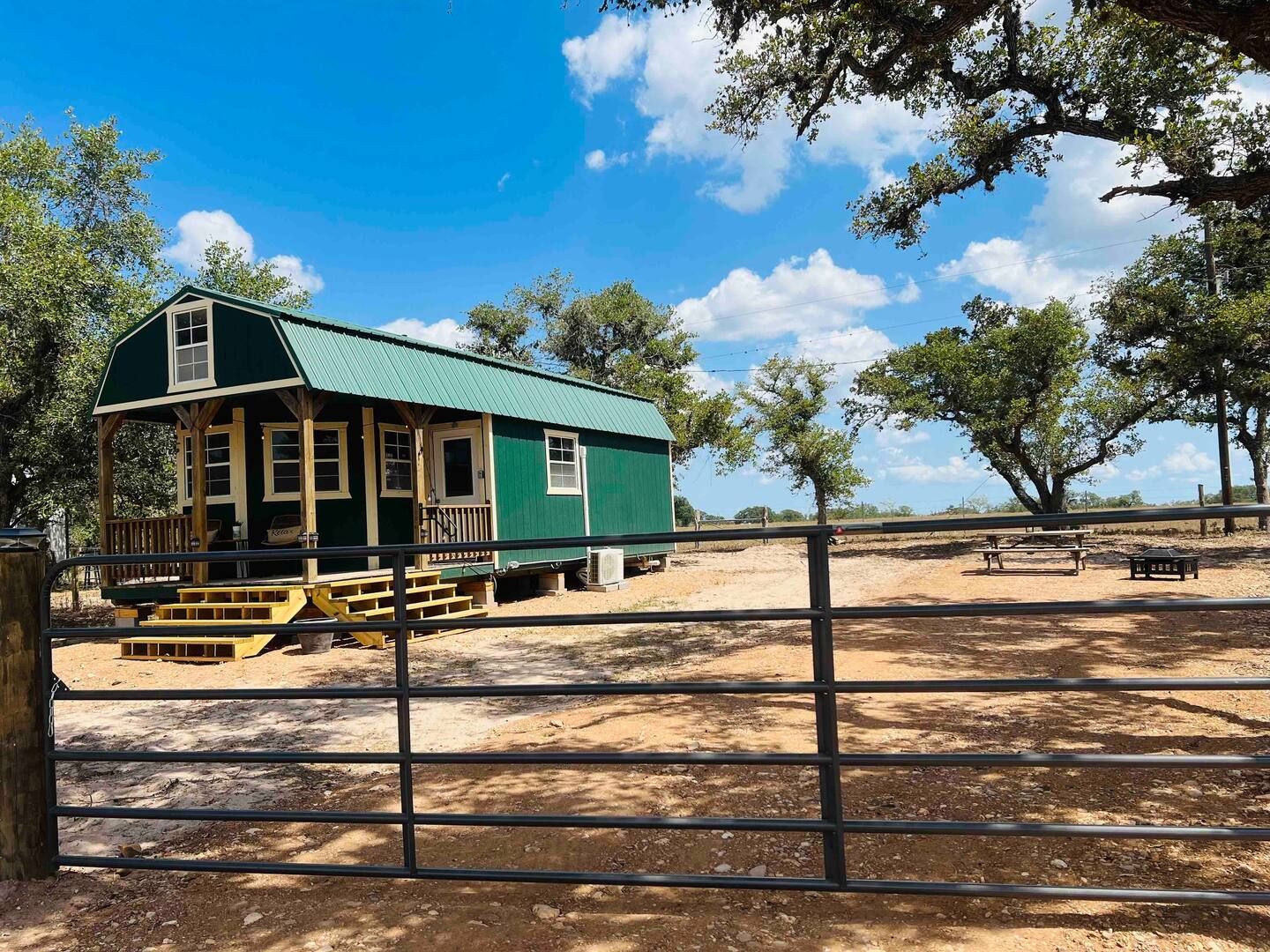 Green farmhouse and porch behind a metal gate in a dry, fenced yard under a blue sky with trees.