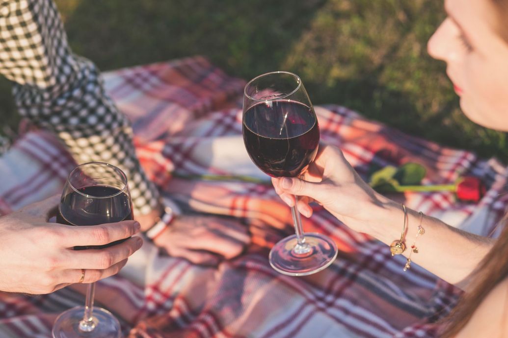 Two people holding wine glasses over a colorful picnic blanket outdoors