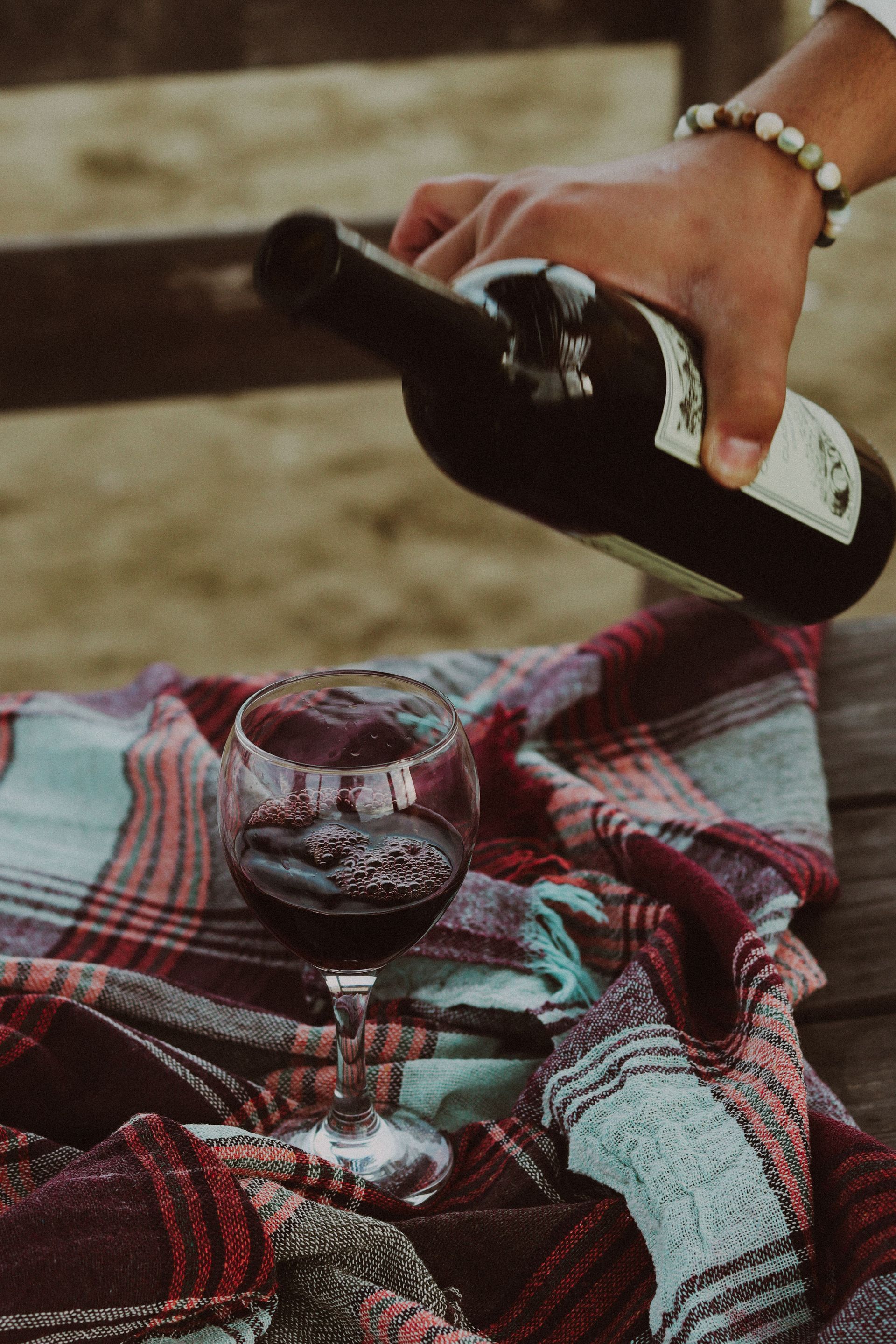Hand pouring red wine into a glass on a plaid blanket outdoors