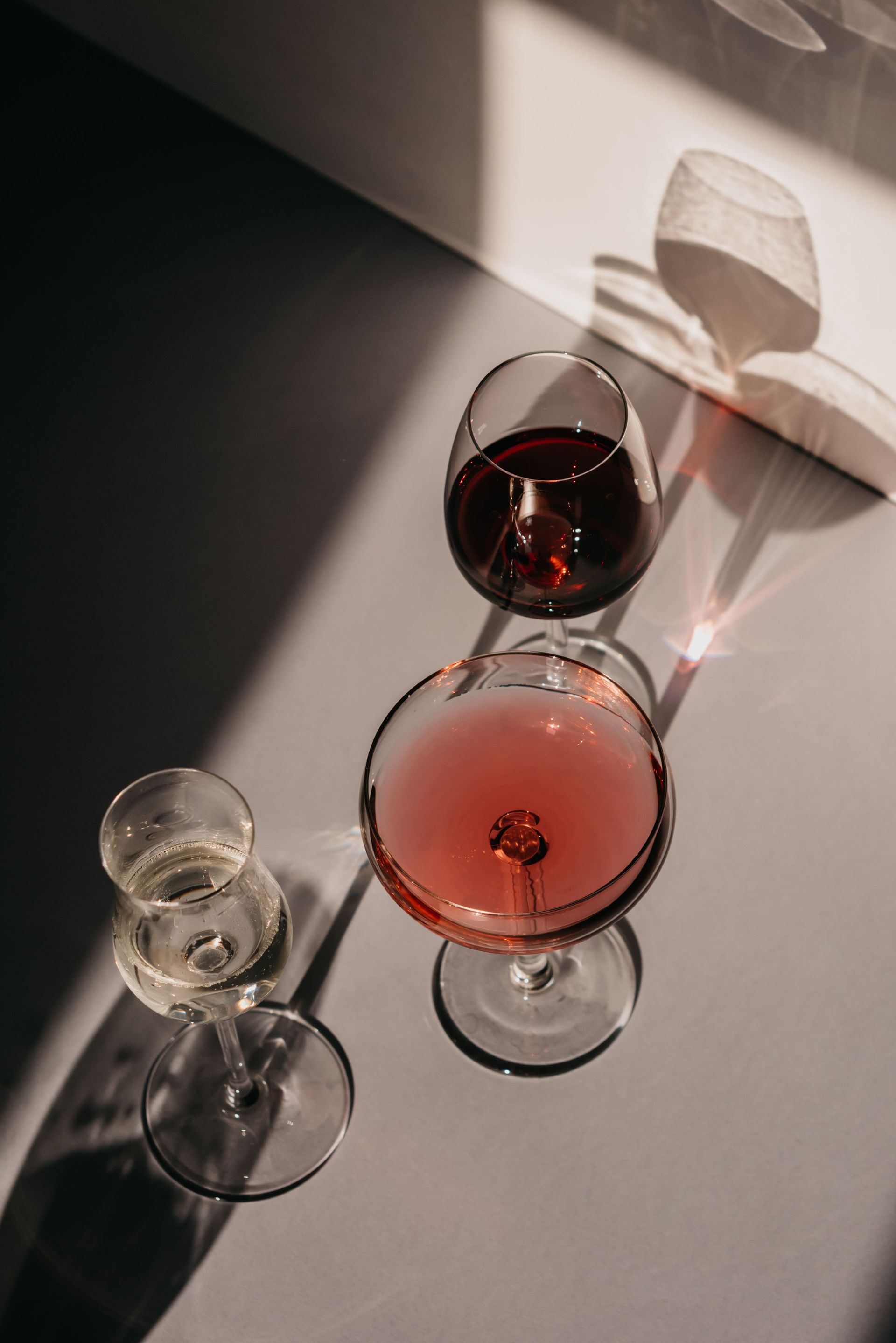 Two wine glasses with red drinks on a sunlit table, casting long shadows.