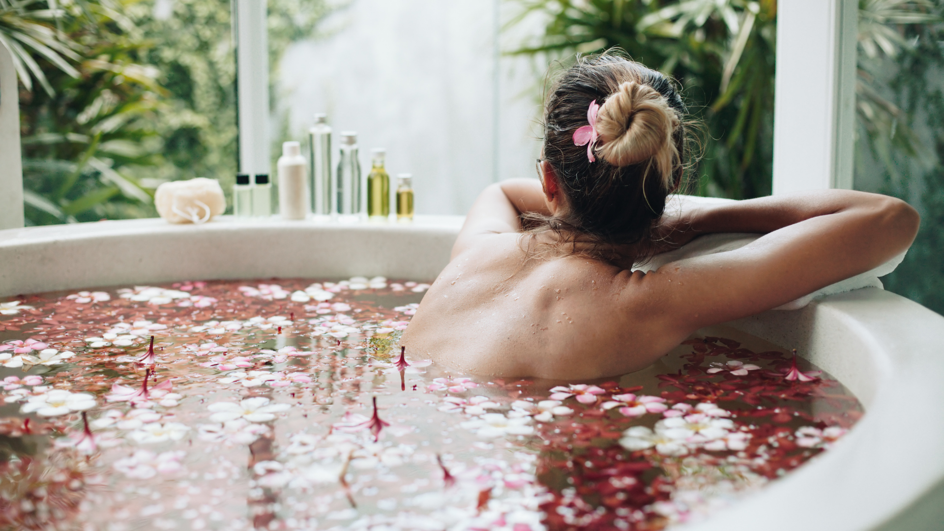 Person relaxing in a rose petal bath, seen from behind, with candles and spa products on the ledge