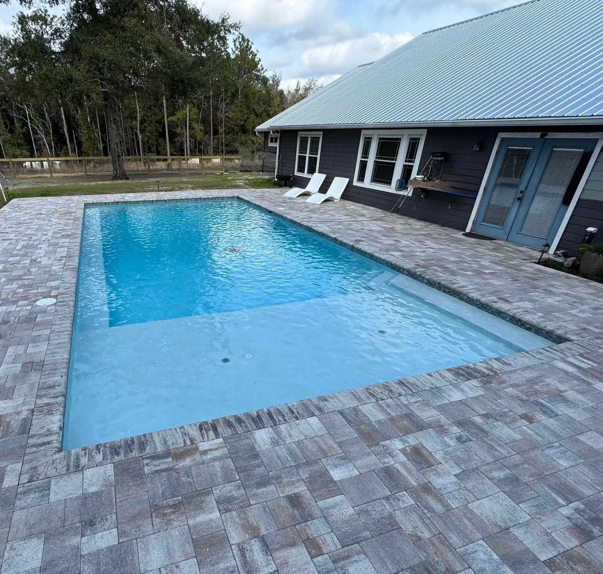 Rectangular backyard swimming pool with blue water beside a brick house and gray paver patio