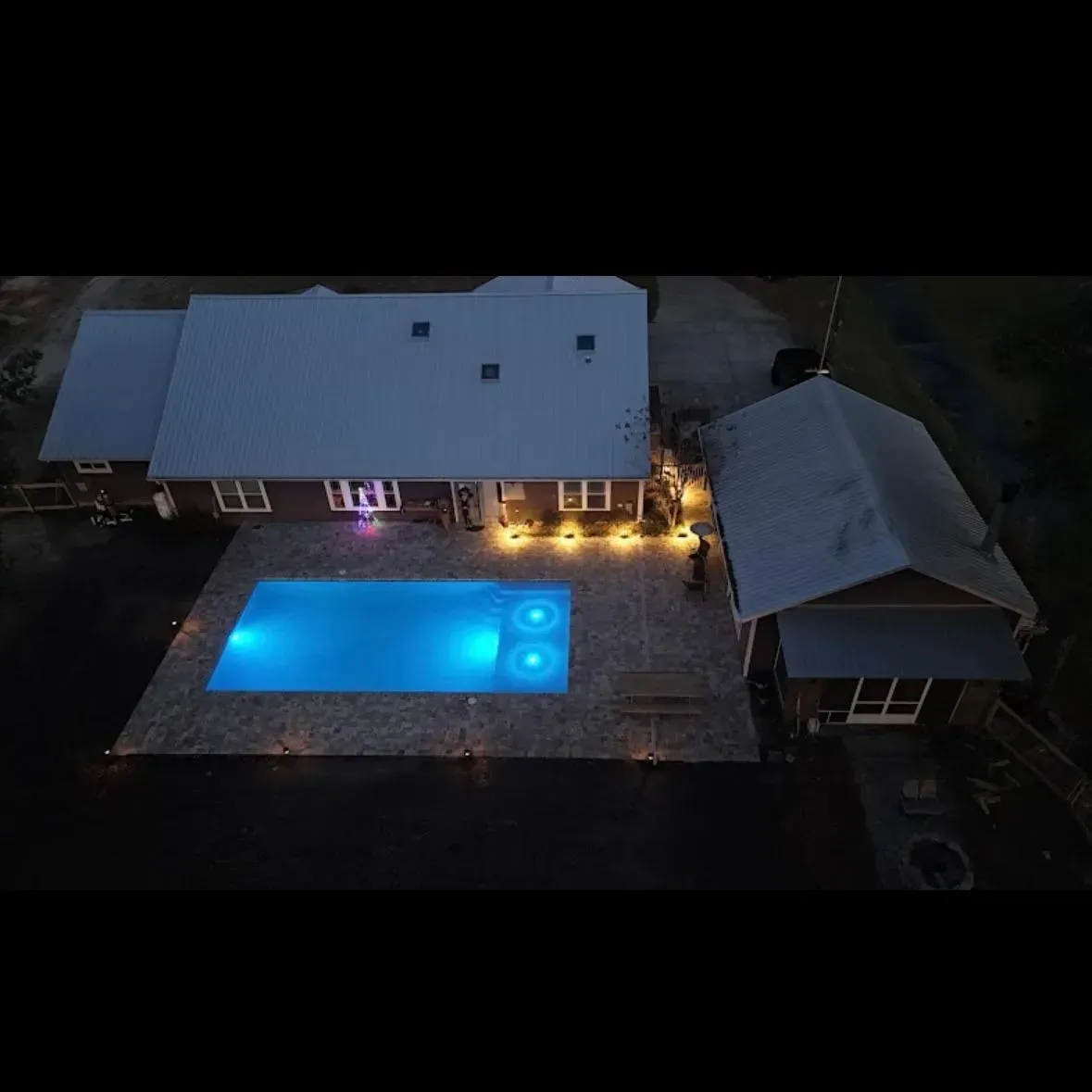 Aerial view of a house at night with a lit blue swimming pool and patio lights.