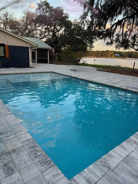 Backyard swimming pool with blue water beside a house, framed by trees at sunset.