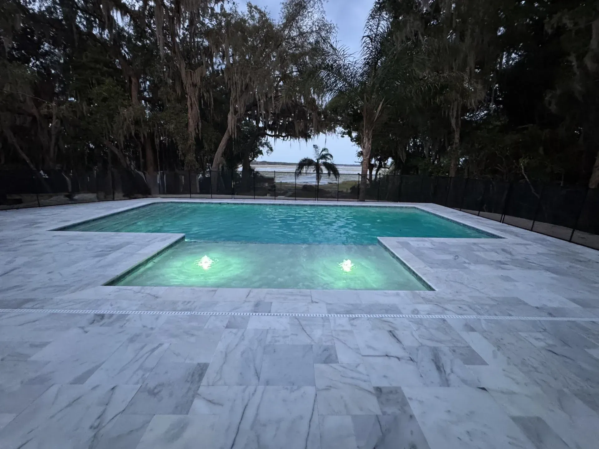Modern backyard pool at dusk with glowing underwater lights, framed by trees and a view of the ocean.