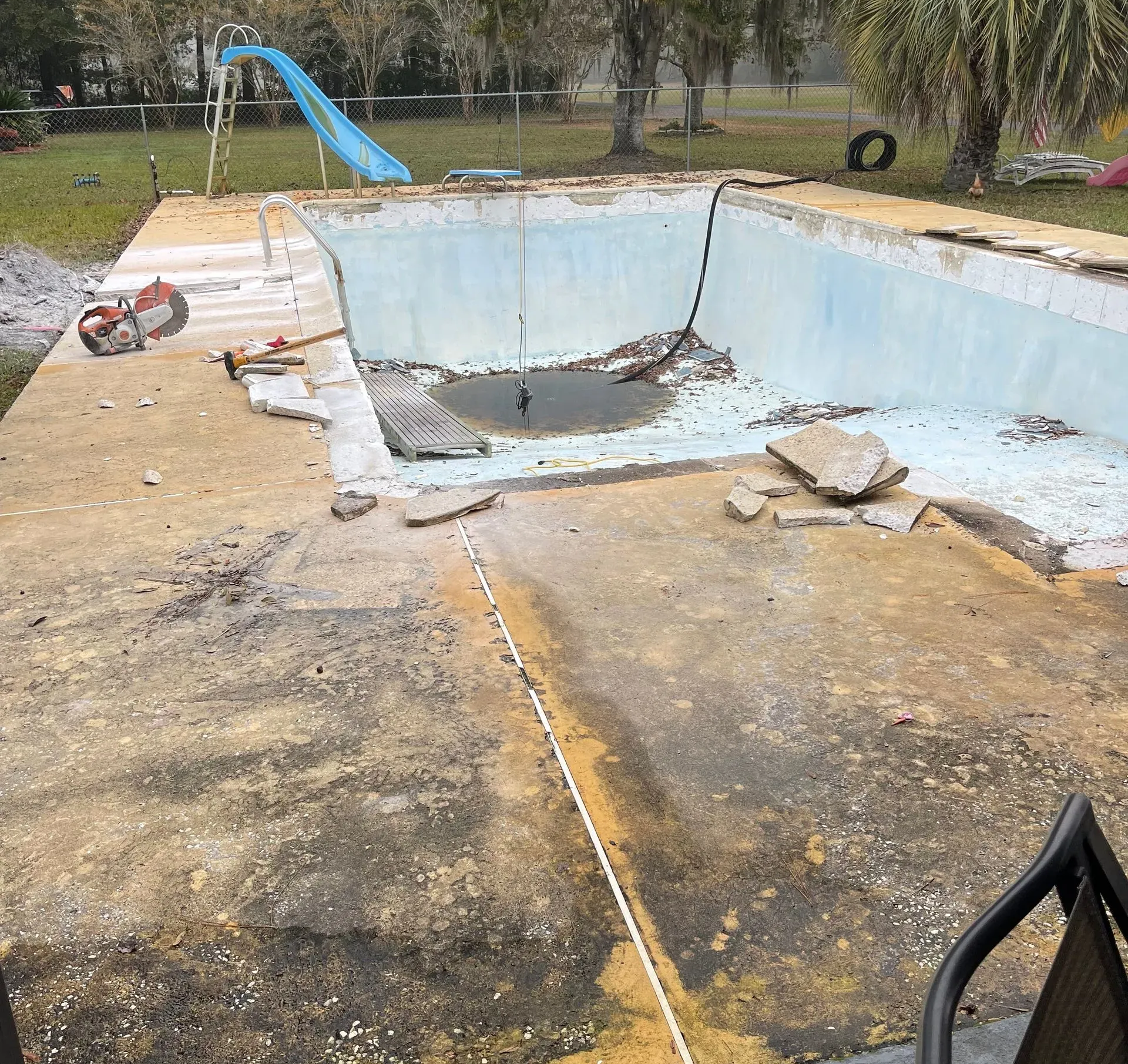 Empty, cracked swimming pool with debris and a blue slide in a backyard.