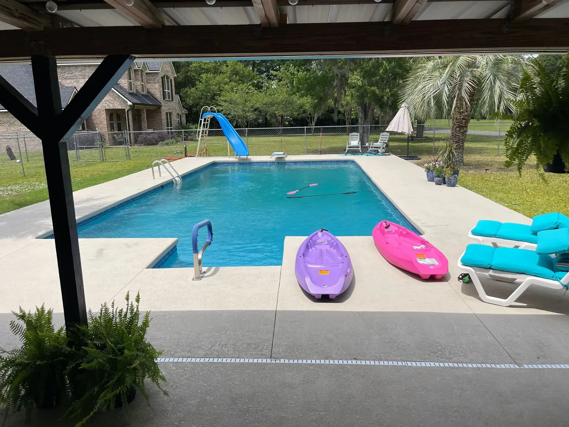 Backyard pool with purple and pink kayaks, blue lounge chairs, and a slide under a shaded patio