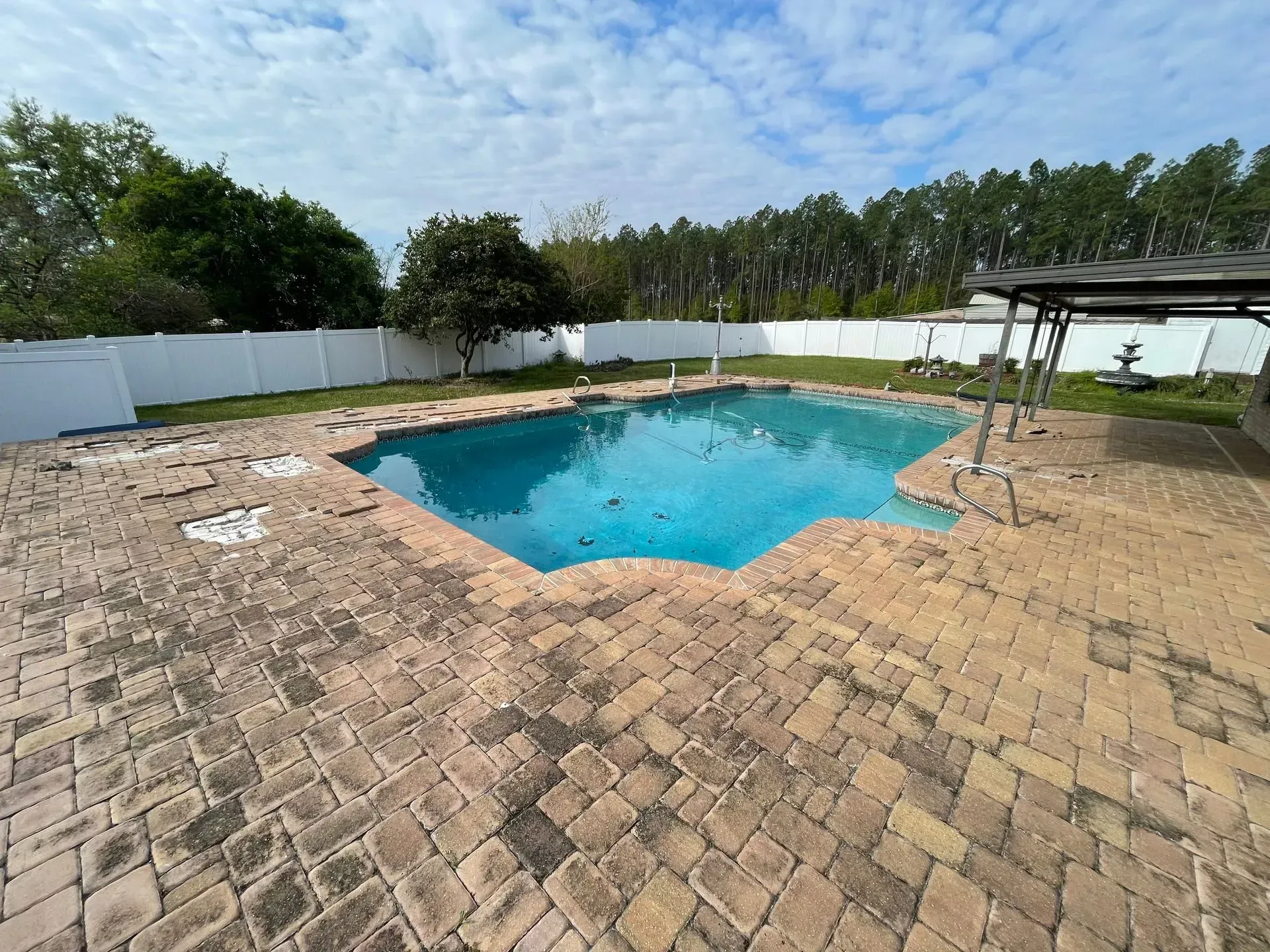 Backyard pool with brick patio, white fence, trees, and a small covered seating area