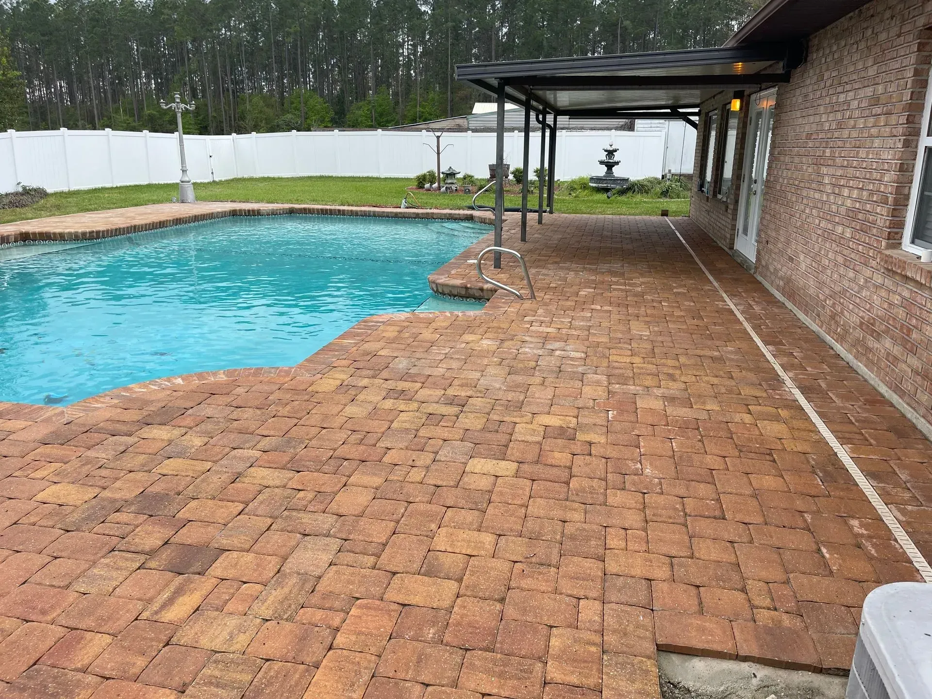 Brick patio beside a turquoise swimming pool under a covered deck, with white fencing and trees beyond.