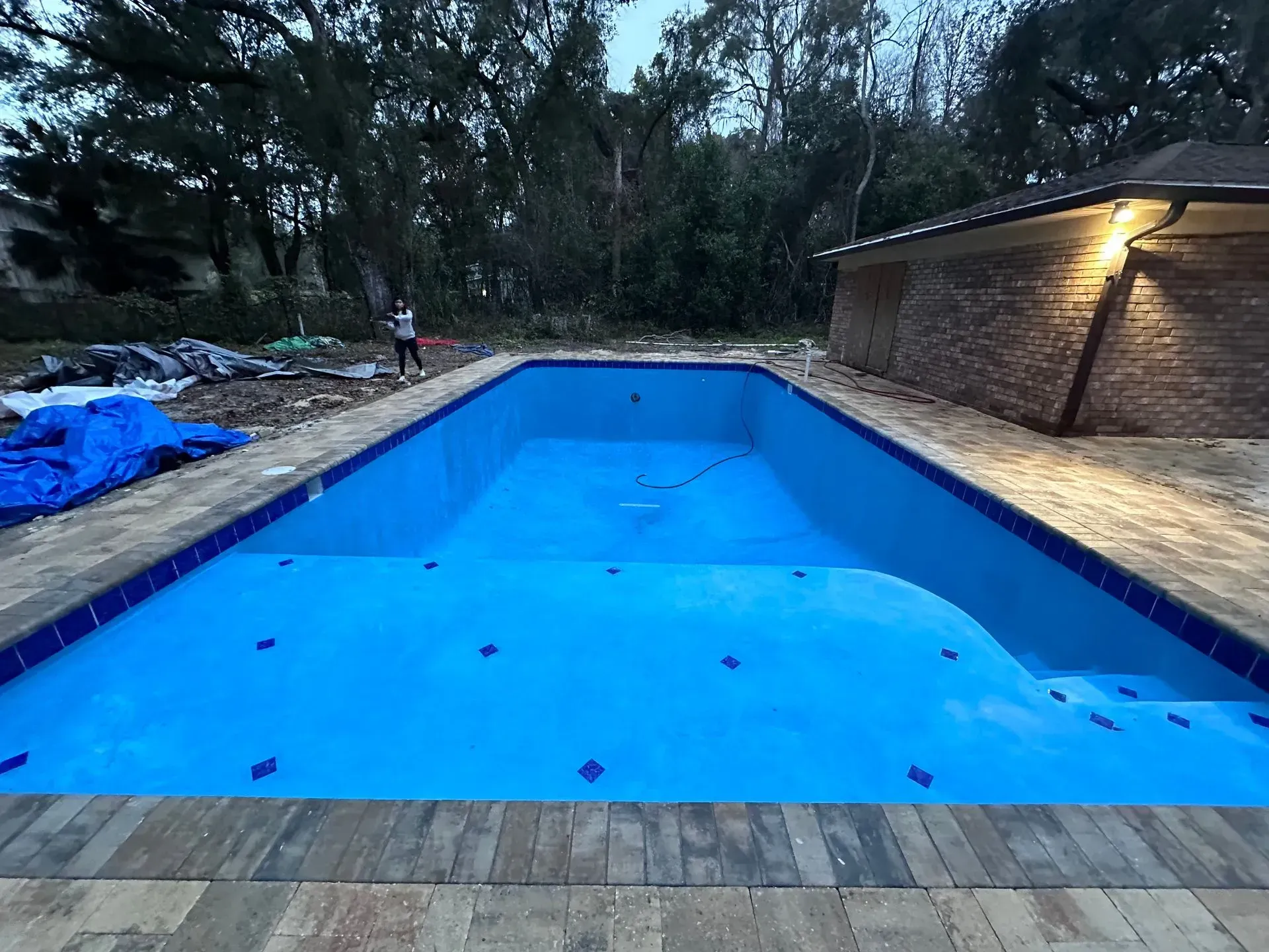 Empty blue swimming pool beside a brick patio at dusk, surrounded by trees and tarps