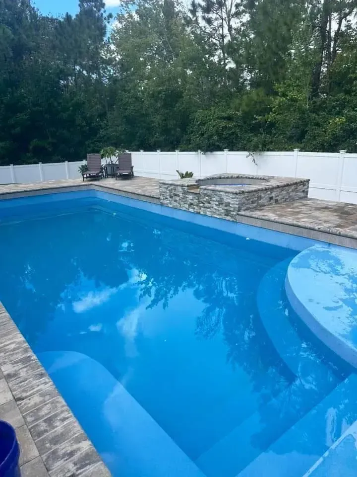 Outdoor backyard swimming pool with bright blue water, stone deck, white fence, and trees in the background