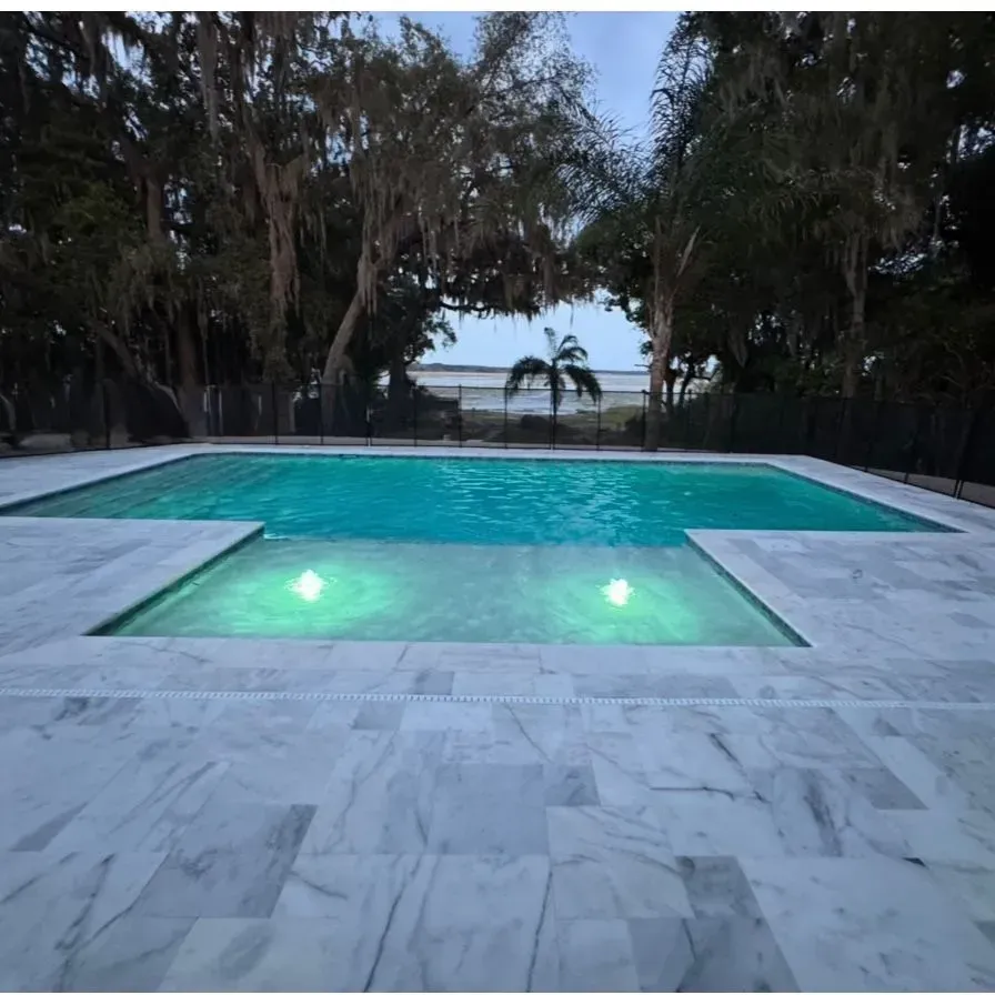 Marble-tiled patio with a lit turquoise pool, framed by trees at dusk, overlooking the water.