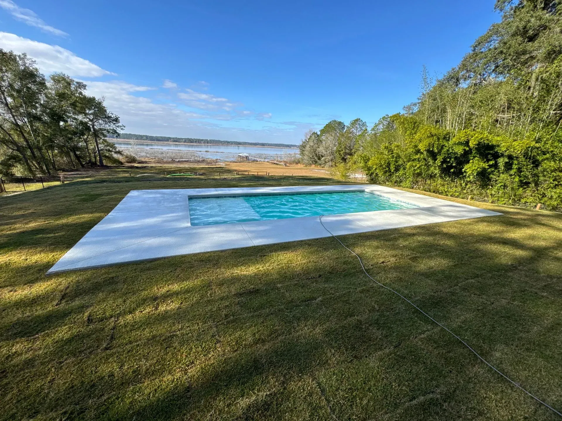 Minimalist white pool deck with a turquoise pool overlooking a grassy field and trees under a blue sky